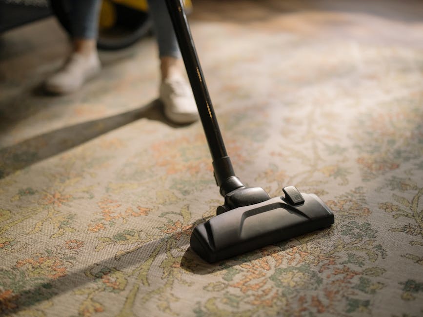 A person kneeling on a patterned carpet in a room, preparing a yellow and black vacuum cleaner for deep cleaning. The individual is opening the vacuum's compartment, with the machine's hose extended o