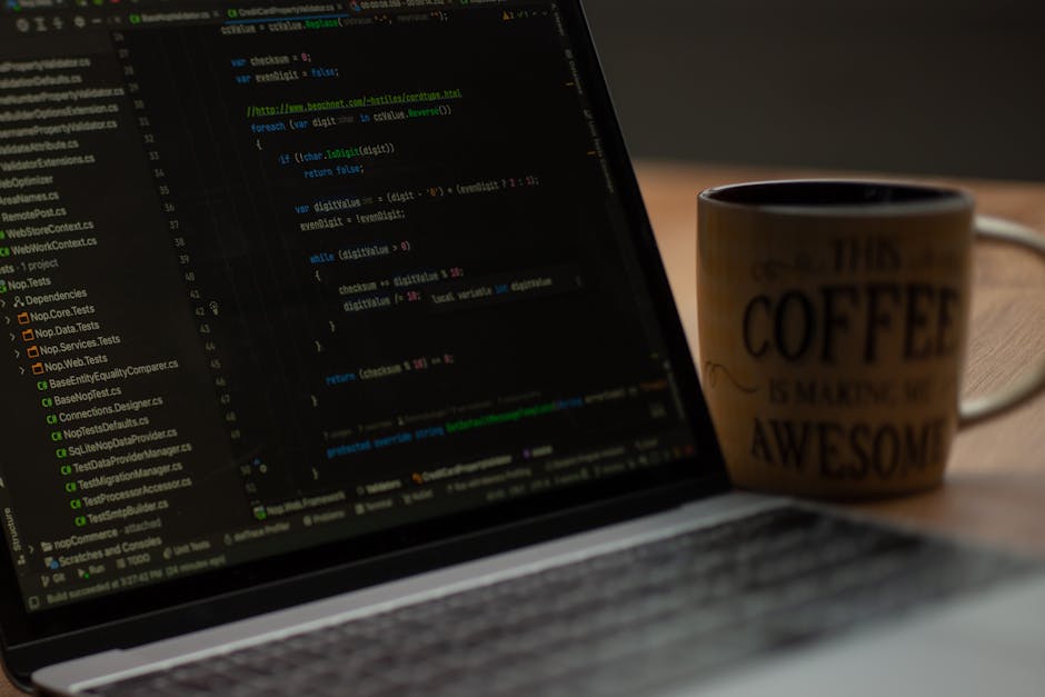 A close-up view of a laptop displaying lines of code on a dark background, positioned on a wooden desk. Next to the laptop is a ceramic mug with text that reads 'THIS COFFEE IS MAKING ME AWESOME,' whi